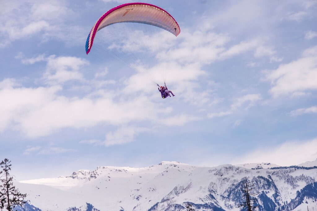 A vibrant paraglider soars through a clear blue sky, with fluffy white clouds dotting the horizon. The paraglider is a bright red and white, with a person seated comfortably inside. In the distance, snow-capped mountains rise majestically, creating a stunning backdrop to this aerial adventure.